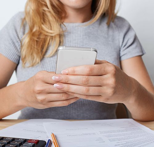 Cropped shot of young female enterpreneur in grey T-shirt, holding smart phone in hands, resting for minute after paper work at office. Businesswoman surfing internet on cell phone during working day