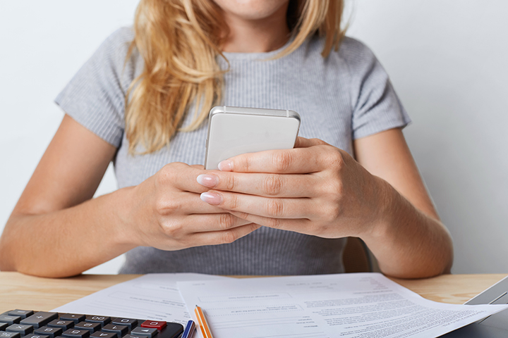 Cropped shot of young female enterpreneur in grey T-shirt, holding smart phone in hands, resting for minute after paper work at office. Businesswoman surfing internet on cell phone during working day